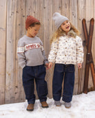 Two children standing in snow in front of a wooden wall, wearing winter clothing. The child on the left wears a gray knit sweater with a red and white 'APRÈS-SJ' stripe, dark blue jeans, and a brown beanie.