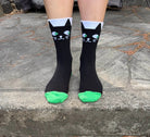 Black crew socks with a cartoon cat face design and bright green toes, worn on feet standing on a stone surface.