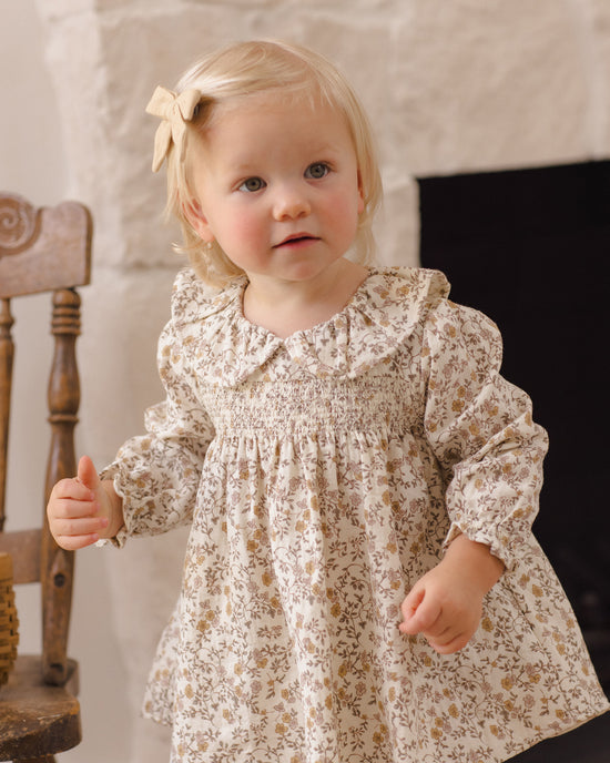 A toddler girl with blonde hair and a bow wearing a long-sleeved cream dress with a small brown floral print, standing in a room with a stone wall and a wooden chair.