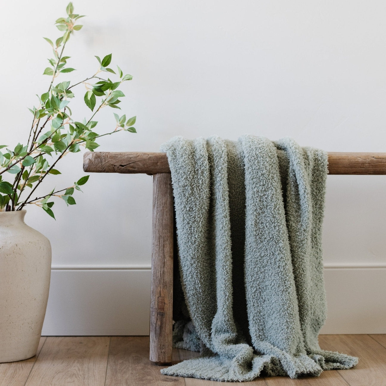 Green textured blanket draped over a wooden bench next to a vase with greenery on a white wall background