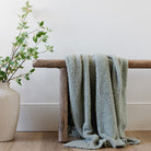 Green textured blanket draped over a wooden bench next to a vase with greenery on a white wall background