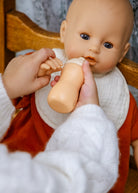 A close-up of a baby feeding doll bottle scene, showing a person's hands holding a beige baby bottle to a doll's mouth. The doll has a bald head, realistic skin-toned face, blue eyes, a white bib, and an orange-red outfit, seated in a wooden chair.