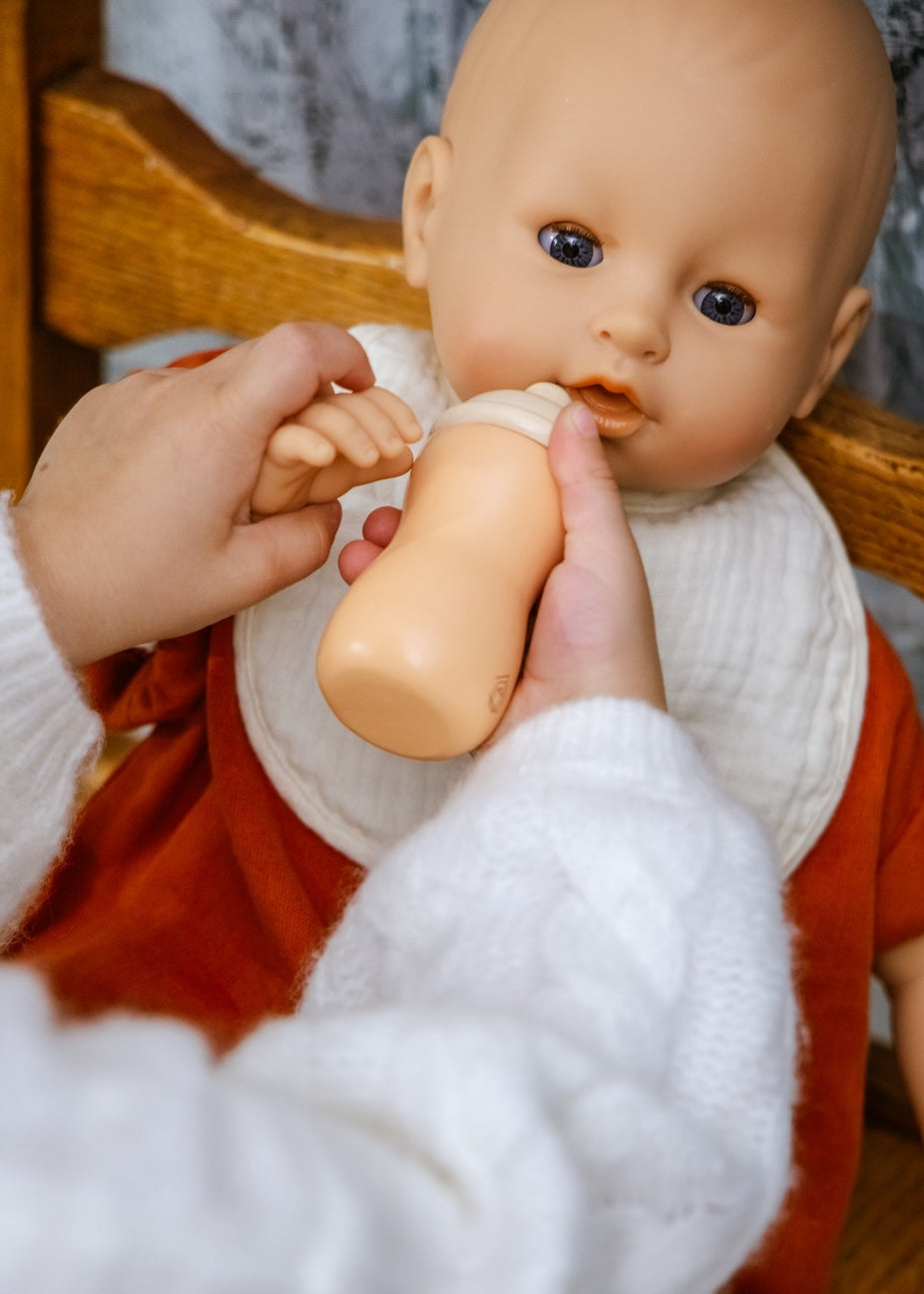 A close-up of a baby feeding doll bottle scene, showing a person's hands holding a beige baby bottle to a doll's mouth. The doll has a bald head, realistic skin-toned face, blue eyes, a white bib, and an orange-red outfit, seated in a wooden chair.