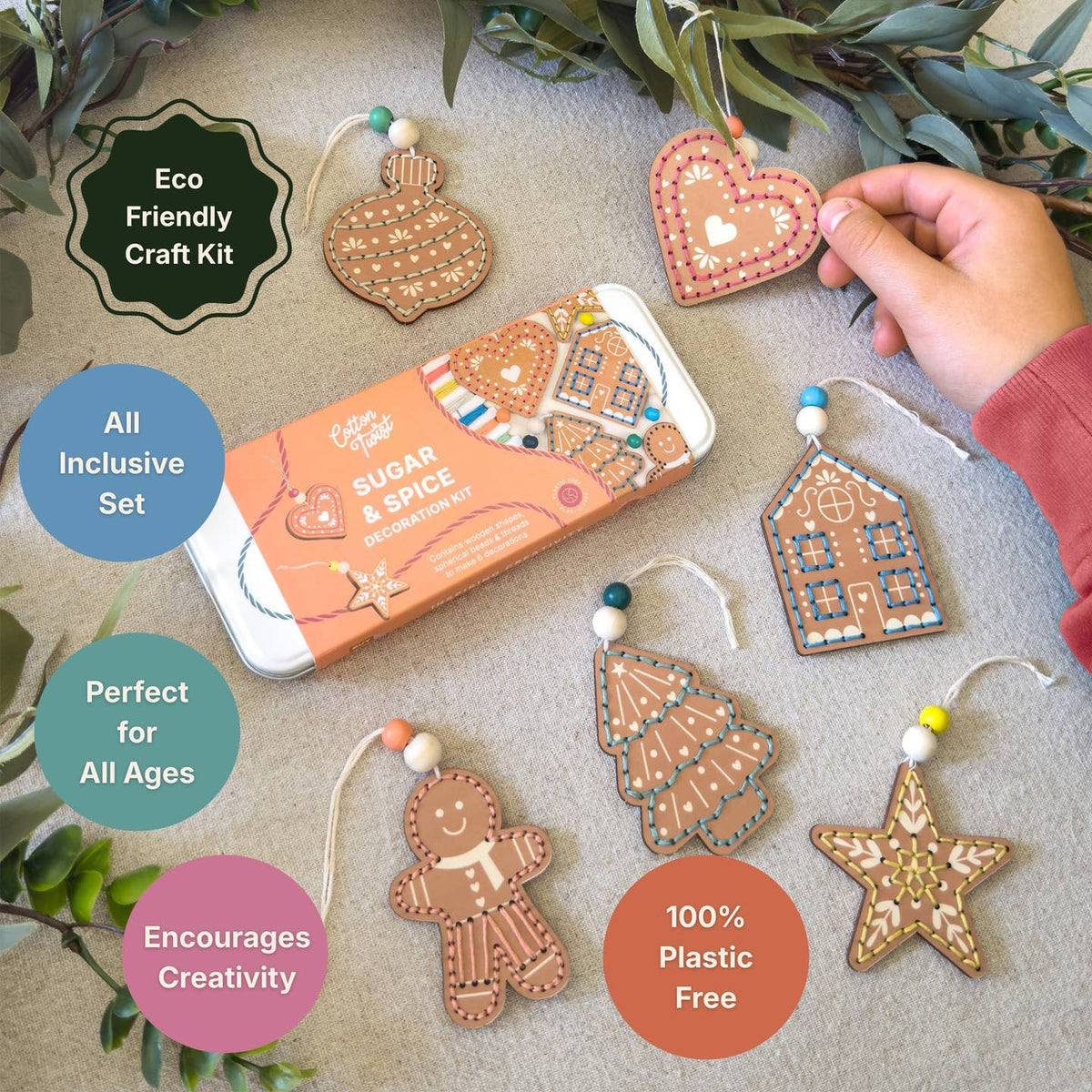 A hand is holding a decorated gingerbread heart Christmas ornament next to a tin of 'Sugar & Spice' eco-friendly craft kit, surrounded by other gingerbread ornaments (tree, house, star, gingerbread man) and promotional text badges on a neutral background.