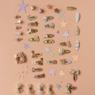 A flat-lay collection of pastel hair clips and decorative hair accessories arranged on a solid peach-pink background. The set includes various star, bow, bunny, and floral hair clips in soft pastel colors like pink, beige, green, and lavender.