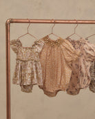 Three baby romper outfits hanging on a copper-colored clothing rack against a textured beige wall. The outfits feature vintage floral and paisley patterns in muted tones of beige, tan, and dusty pink.