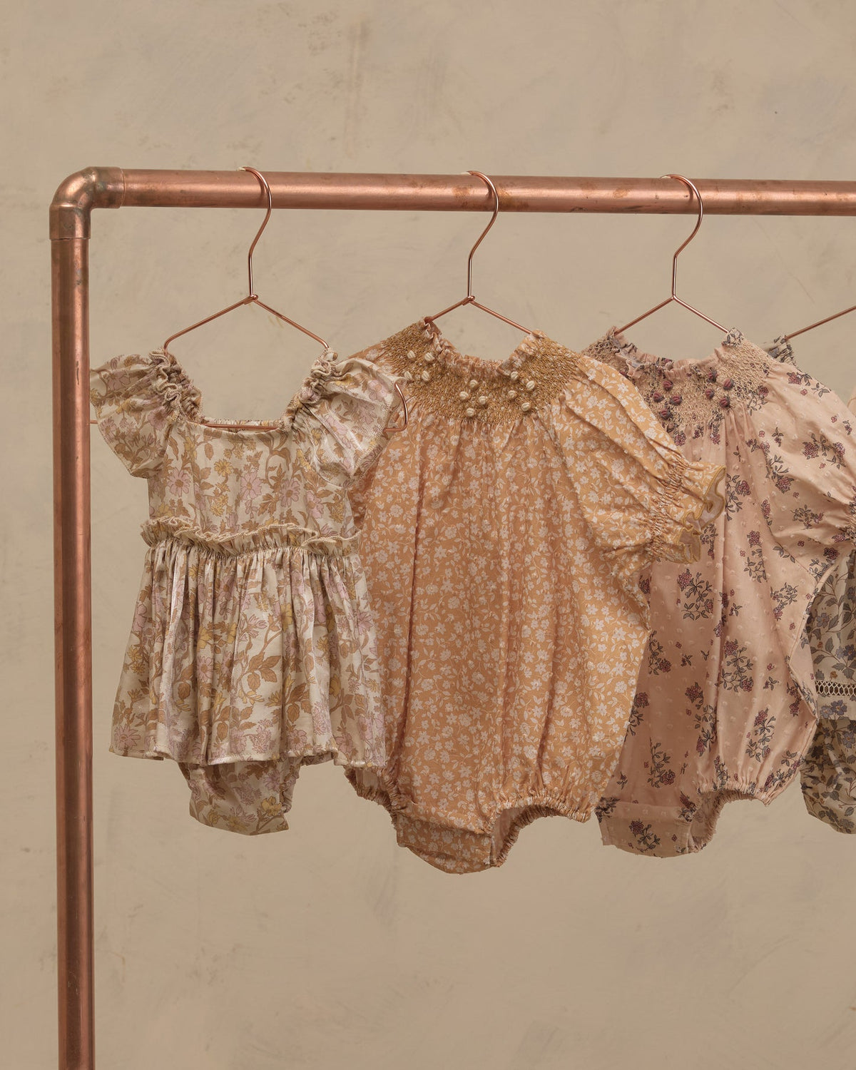 Three baby romper outfits hanging on a copper-colored clothing rack against a textured beige wall. The outfits feature vintage floral and paisley patterns in muted tones of beige, tan, and dusty pink.