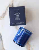 A blue glass jar candle and its dark navy Botanica 'Après Ski' packaging box, styled on a white marble surface.