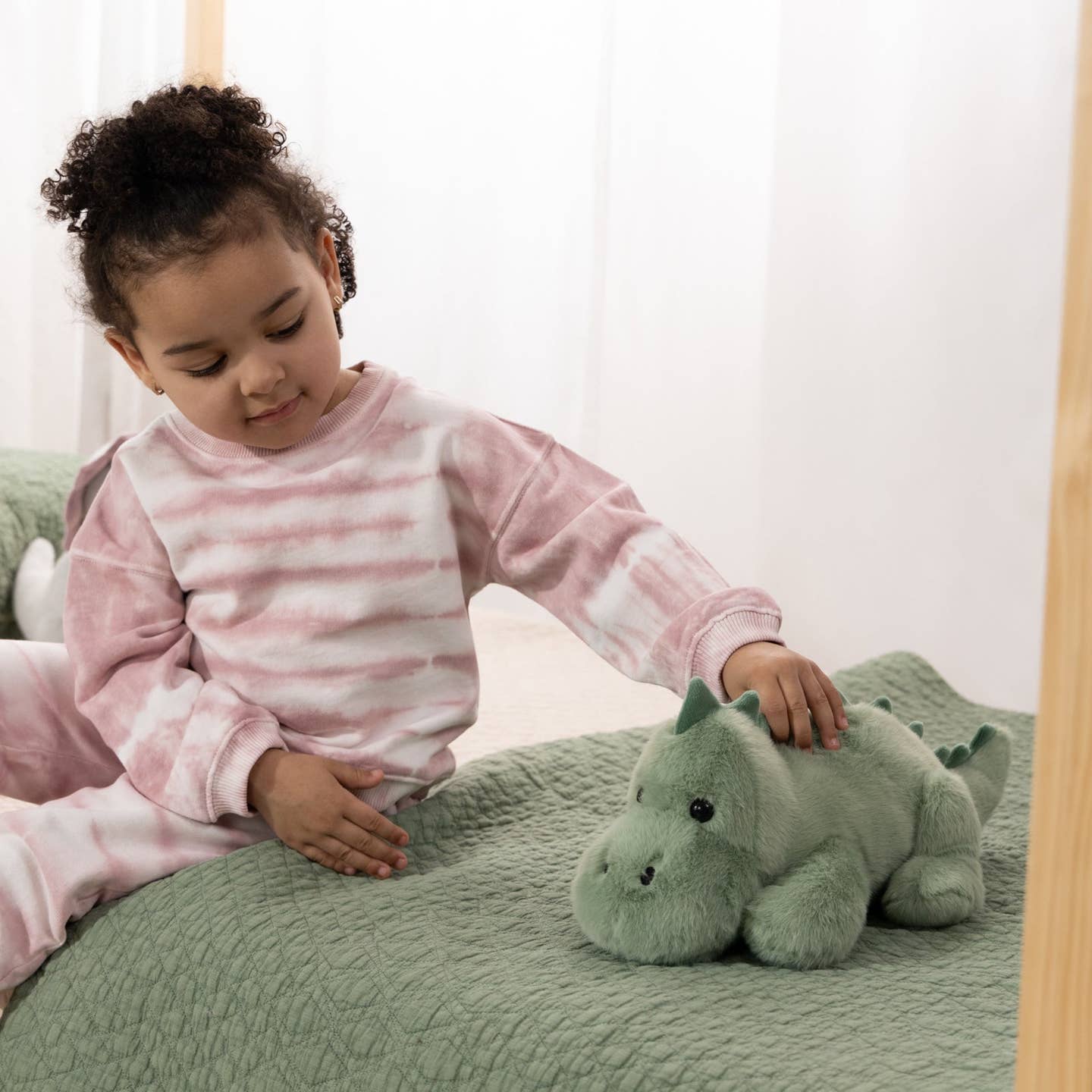 A young child wearing pink and white striped pajamas sits on a bed, gently touching a small green plush dinosaur toy.