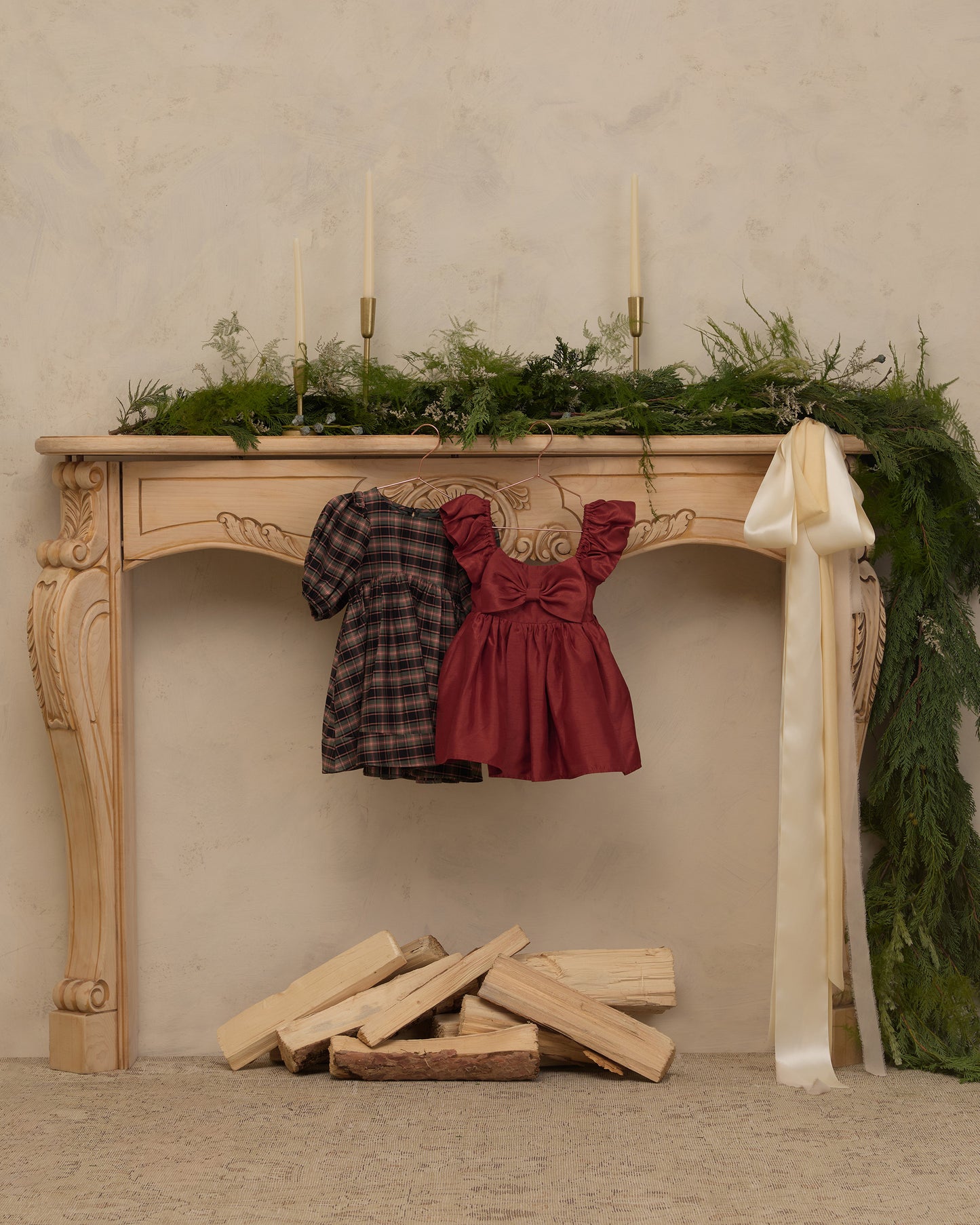 Two children's dresses hanging from an ornate wooden console table in a styled indoor scene. The left dress is dark plaid, and the right dress is a dusty rose red ruffled style.