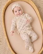 A baby wearing a cream-colored footed sleeper with a matching bonnet, decorated with tiny brown cross patterns, lying on a beige cushion inside a woven oval basket.