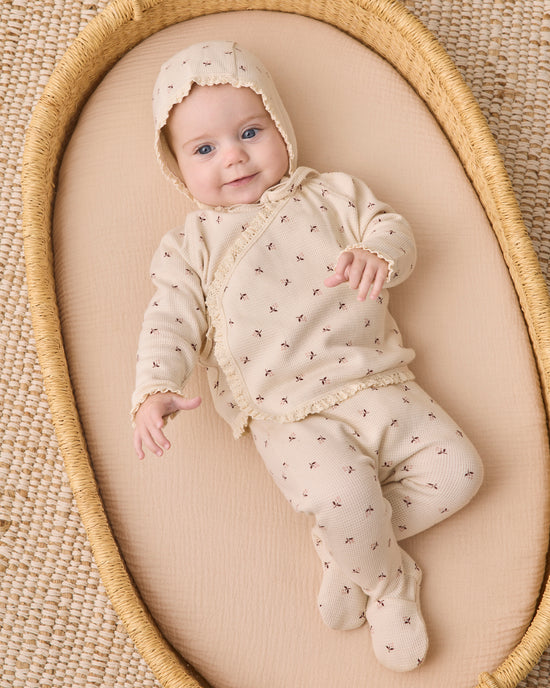 A baby wearing a cream-colored footed sleeper with a matching bonnet, decorated with tiny brown cross patterns, lying on a beige cushion inside a woven oval basket.