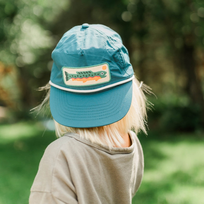 A child seen from behind wearing a teal blue cap with a fish patch on the front, standing outdoors in a grassy, tree-filled area.