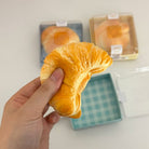 Hand holding a soft, yellow bread-like toy with more toys and a box in the background.