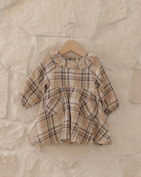 A toddler girl's long-sleeve plaid dress in beige, brown, and black, hanging on a wooden hanger against a rustic stone wall.