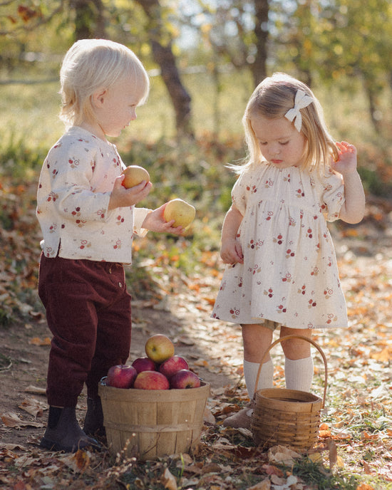 Two young children outdoors in a wooded, autumn setting, wearing matching cream floral outfits. One child holds an apple near a basket of apples, while the other stands nearby with a basket on the ground.