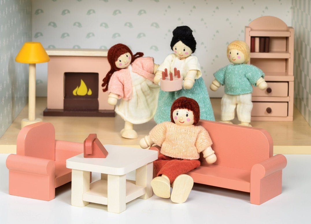 A miniature wooden dollhouse room interior featuring a wooden toy figure standing next to a coral-pink sofa and armchair set, a coffee table with a tiny red telephone, a pale pink decorative fireplace, a yellow floor lamp, and a natural wood bookcase against a patterned wallpaper background.