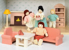 A miniature wooden dollhouse room interior featuring a wooden toy figure standing next to a coral-pink sofa and armchair set, a coffee table with a tiny red telephone, a pale pink decorative fireplace, a yellow floor lamp, and a natural wood bookcase against a patterned wallpaper background.