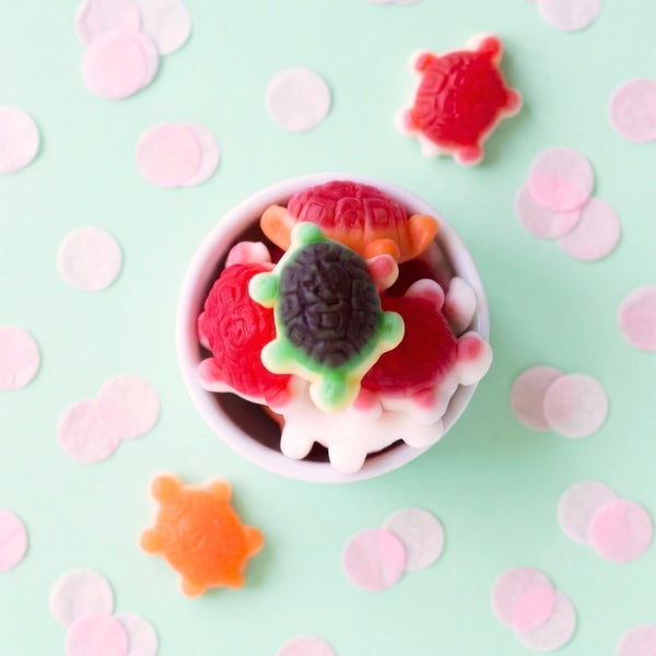 Colorful gummy turtle candies in a bowl on a light green background with pink confetti.