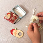 A person's hands are shown wrapping cream-colored yarn around a small wooden spool. On the light beige fabric surface next to them are a red and silver tin of 'Jolly Santa' yarn gift set and a Santa Claus-shaped wooden ornament.