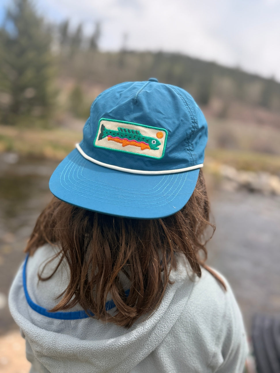 A person viewed from behind wearing a bright blue flat-brim cap outdoors near a stream, with a fish patch on the front and a light gray hoodie.