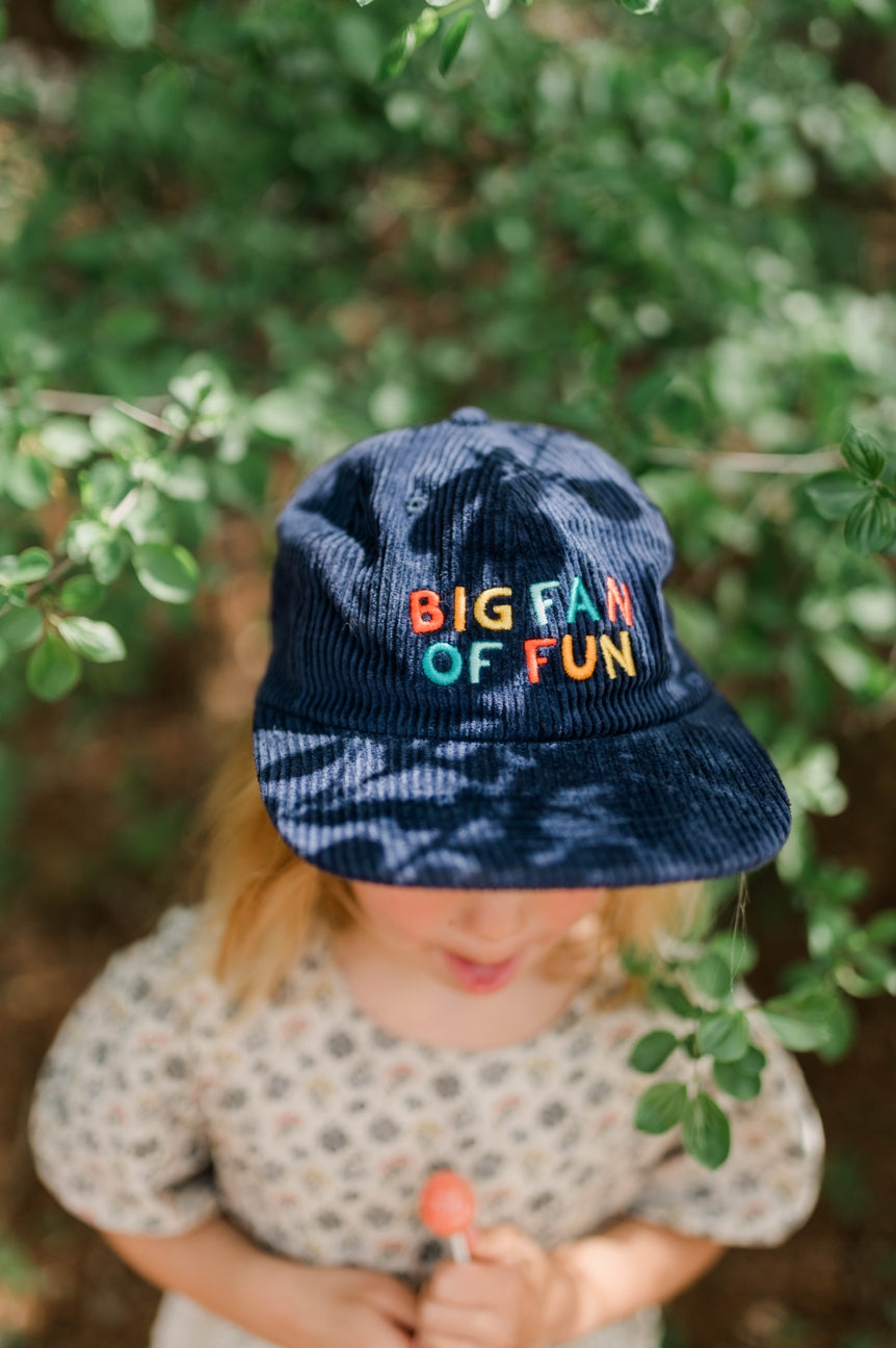 Child wearing a blue cap with colorful text, standing in front of green foliage.