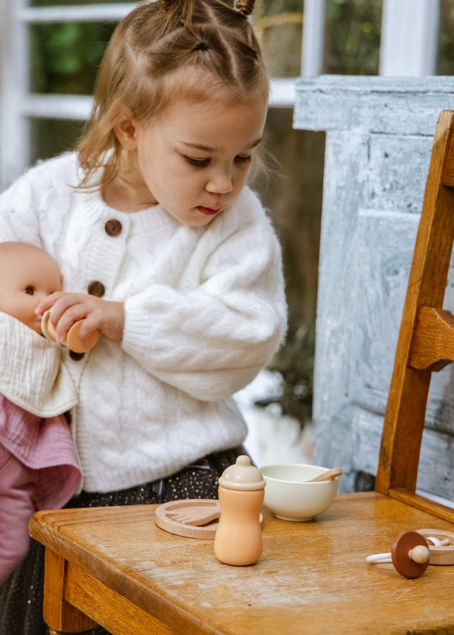 A young child in a white knit cardigan holds a doll beside a wooden table with a wooden toy bottle, bowl, and pacifier in a softly lit indoor setting.