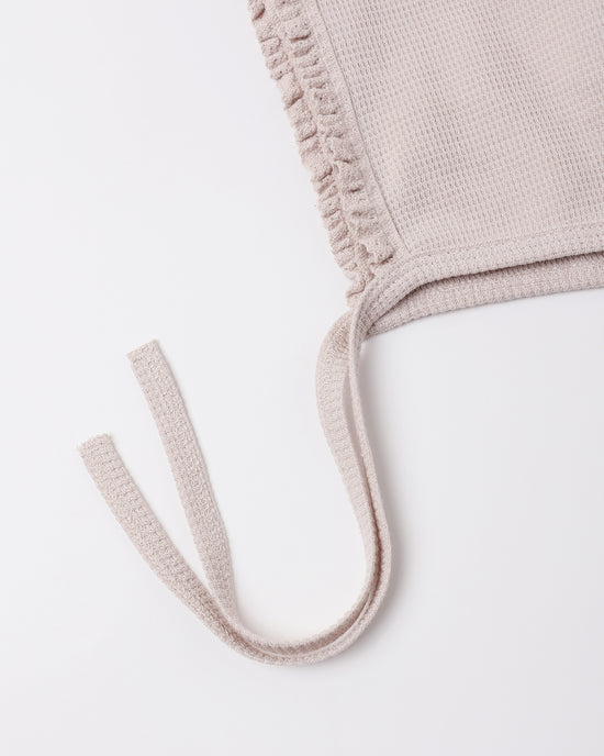 Close-up of a light beige knit garment with a ruffled trim and matching tie straps on a plain white background.