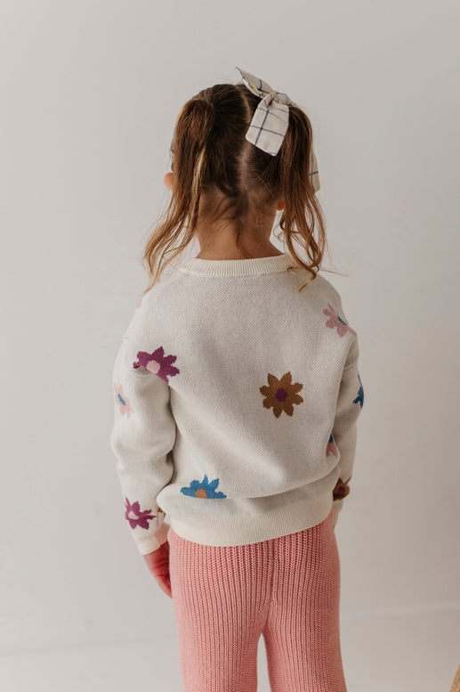 Child wearing a white sweater with colorful flower patterns and pink pants on a plain background