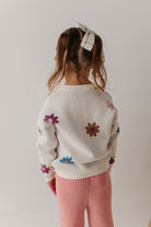 Child wearing a white sweater with colorful flower patterns and pink pants on a plain background