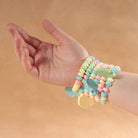 A close-up of a person's hand and wrist wearing a stack of pastel candy-themed bracelets, including pastel beads and elephant-shaped charms, against a solid tan background.