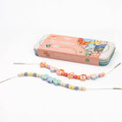 A bead串ing kit with wooden beads, needles, and a pink metal tin box on a white background.