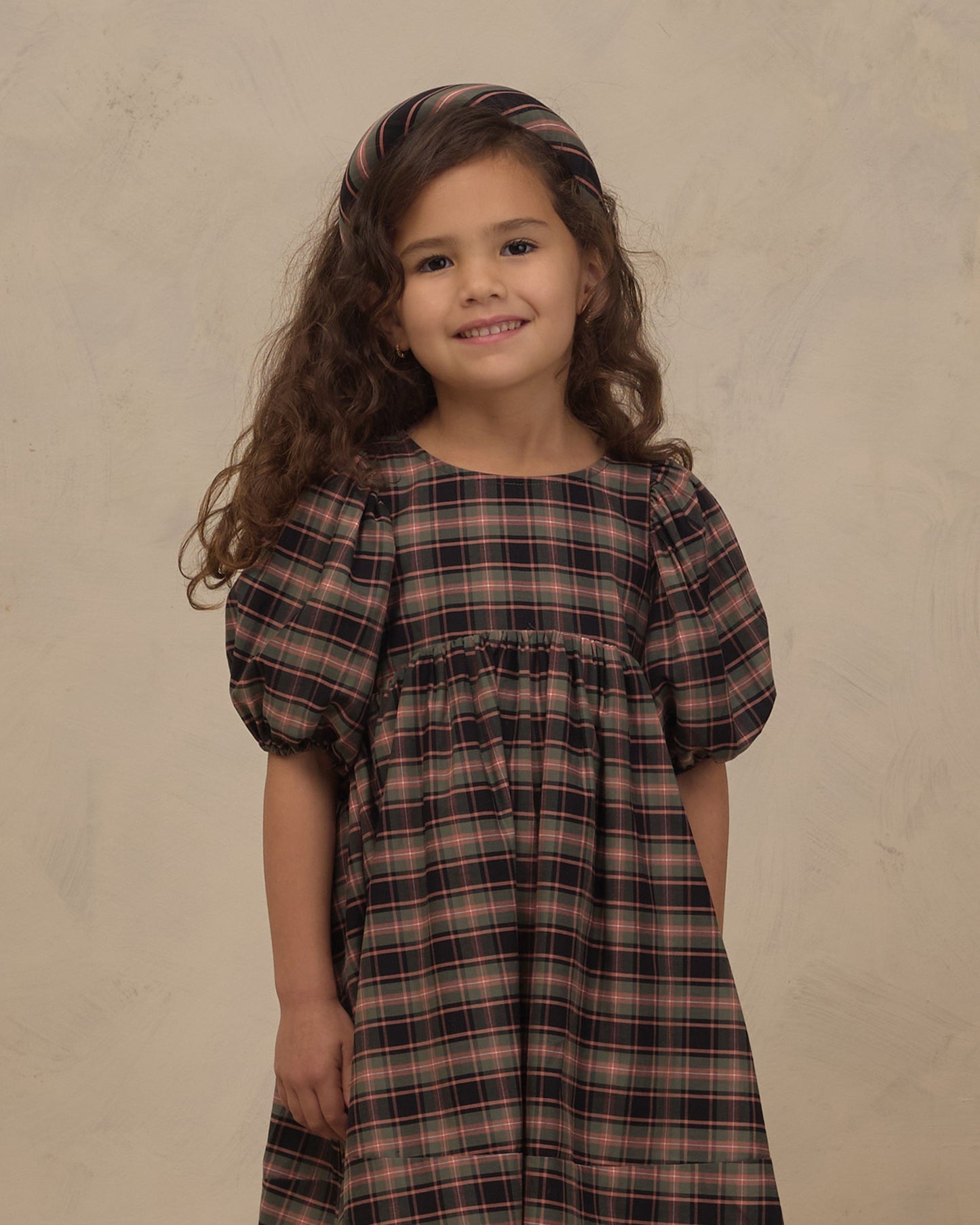 A young girl with long, wavy brown hair wearing a dark brown and black plaid dress with puff sleeves and a matching headband, standing against a textured beige background.