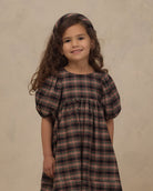 A young girl with long, wavy brown hair wearing a dark brown and black plaid dress with puff sleeves and a matching headband, standing against a textured beige background.