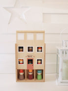 A wooden toy hotel set with a light natural wood facade, six windowed floors, and several small peg-doll characters in colorful clothing displayed inside and on the ground floor. The set is photographed on a white surface against a white paneled wall with a decorative white star and a lantern visible.