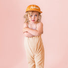 Child wearing a yellow 'Rascal' cap and beige romper against a pink background