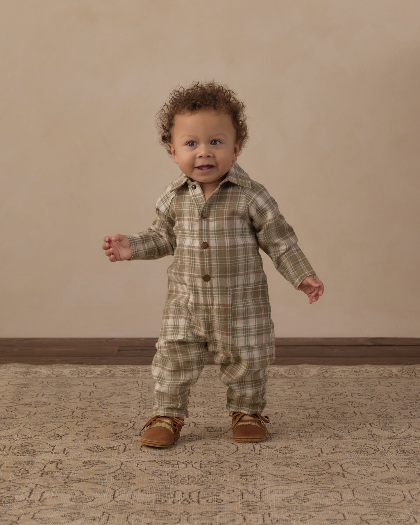 A toddler standing and wearing a long-sleeve plaid romper in muted green, beige, and brown tones, paired with brown shoes, against a warm beige studio backdrop.
