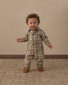 A toddler standing and wearing a long-sleeve plaid romper in muted green, beige, and brown tones, paired with brown shoes, against a warm beige studio backdrop.