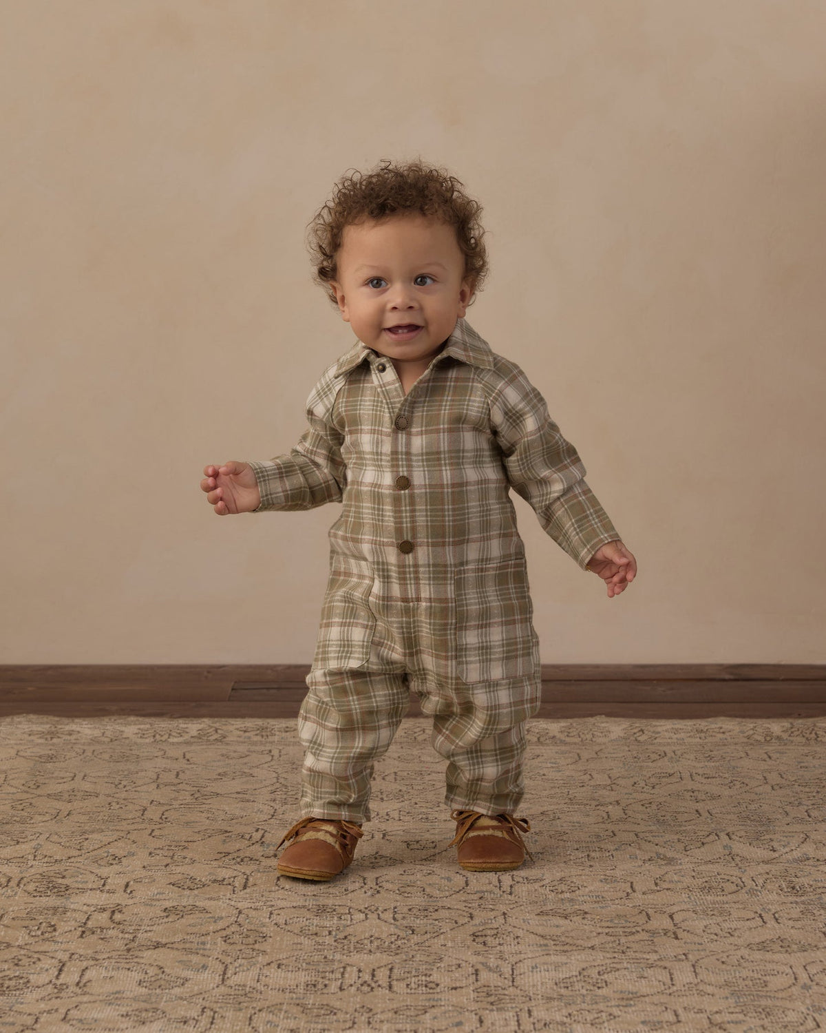 A toddler standing and wearing a long-sleeve plaid romper in muted green, beige, and brown tones, paired with brown shoes, against a warm beige studio backdrop.