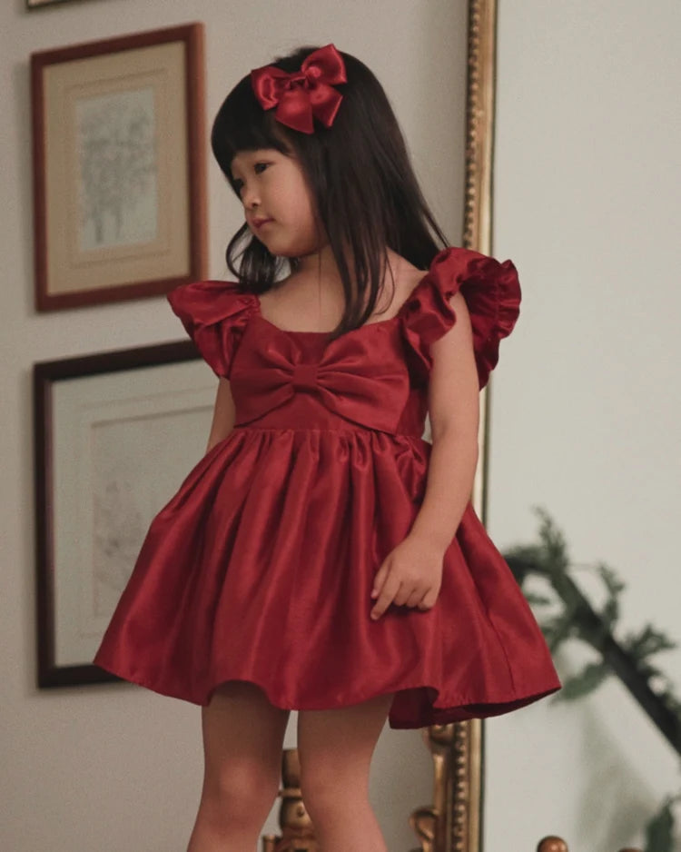 A young girl wearing a shiny deep red off-the-shoulder dress with a large bow at the neckline and a matching red bow headband, standing indoors in front of a mirror.