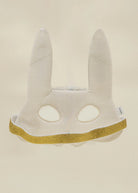 A white bunny mask with long ears and eye cutouts, featuring a textured vertical stripe pattern and a glittery gold trim along the bottom edge, set against a solid beige background.