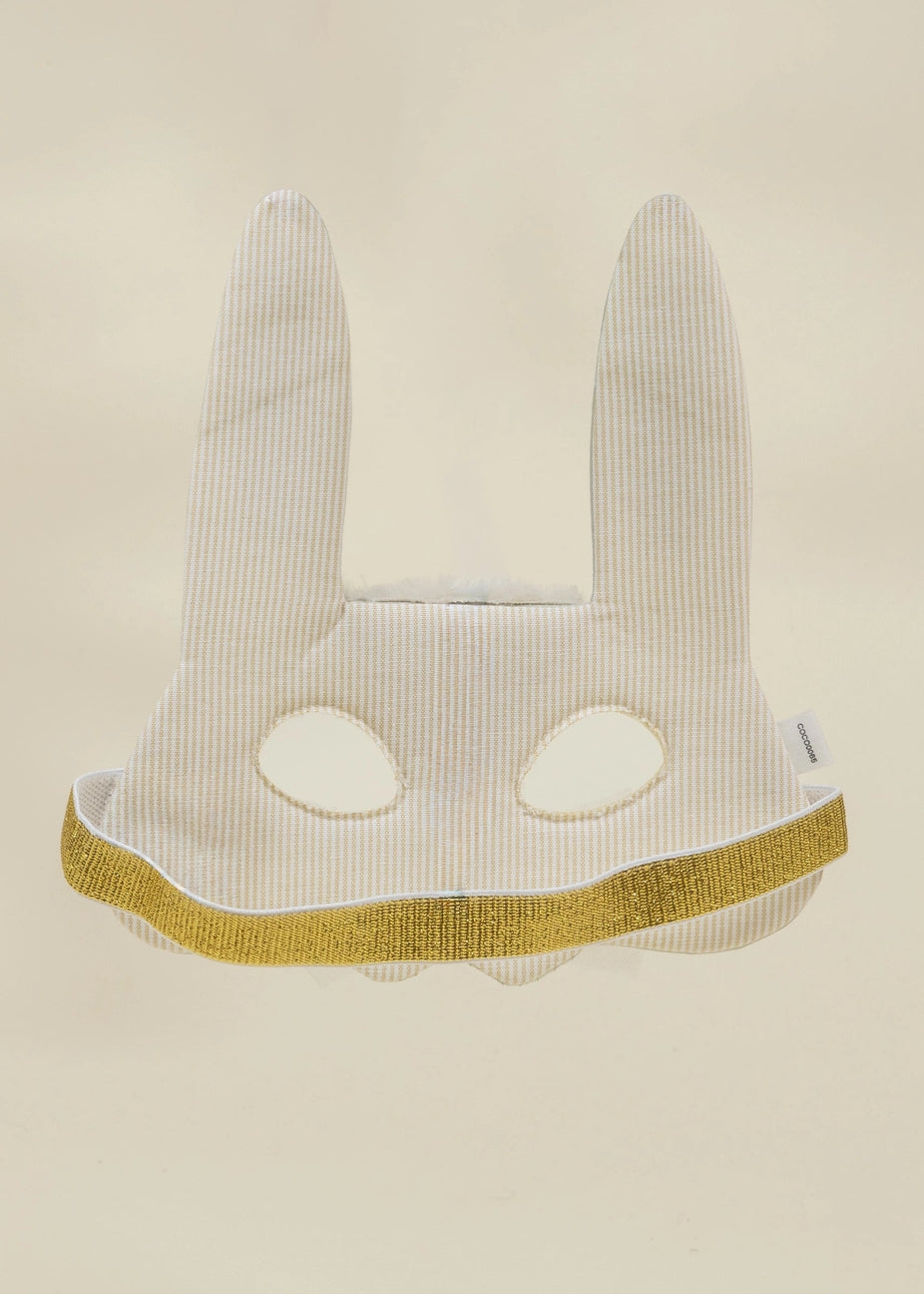A white bunny mask with long ears and eye cutouts, featuring a textured vertical stripe pattern and a glittery gold trim along the bottom edge, set against a solid beige background.