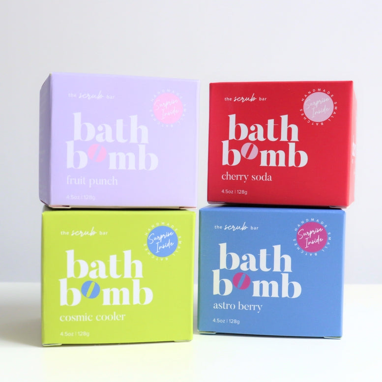 Four boxes of bath bomb by The Scrub Bar stacked in a 2x2 grid on a white surface against a light gray background. The boxes are lavender (fruit punch), red (cherry soda), lime green (cosmic cooler), and blue (astro berry), each with large white text and a small pink circular badge.
