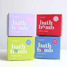 Four boxes of bath bomb by The Scrub Bar stacked in a 2x2 grid on a white surface against a light gray background. The boxes are lavender (fruit punch), red (cherry soda), lime green (cosmic cooler), and blue (astro berry), each with large white text and a small pink circular badge.