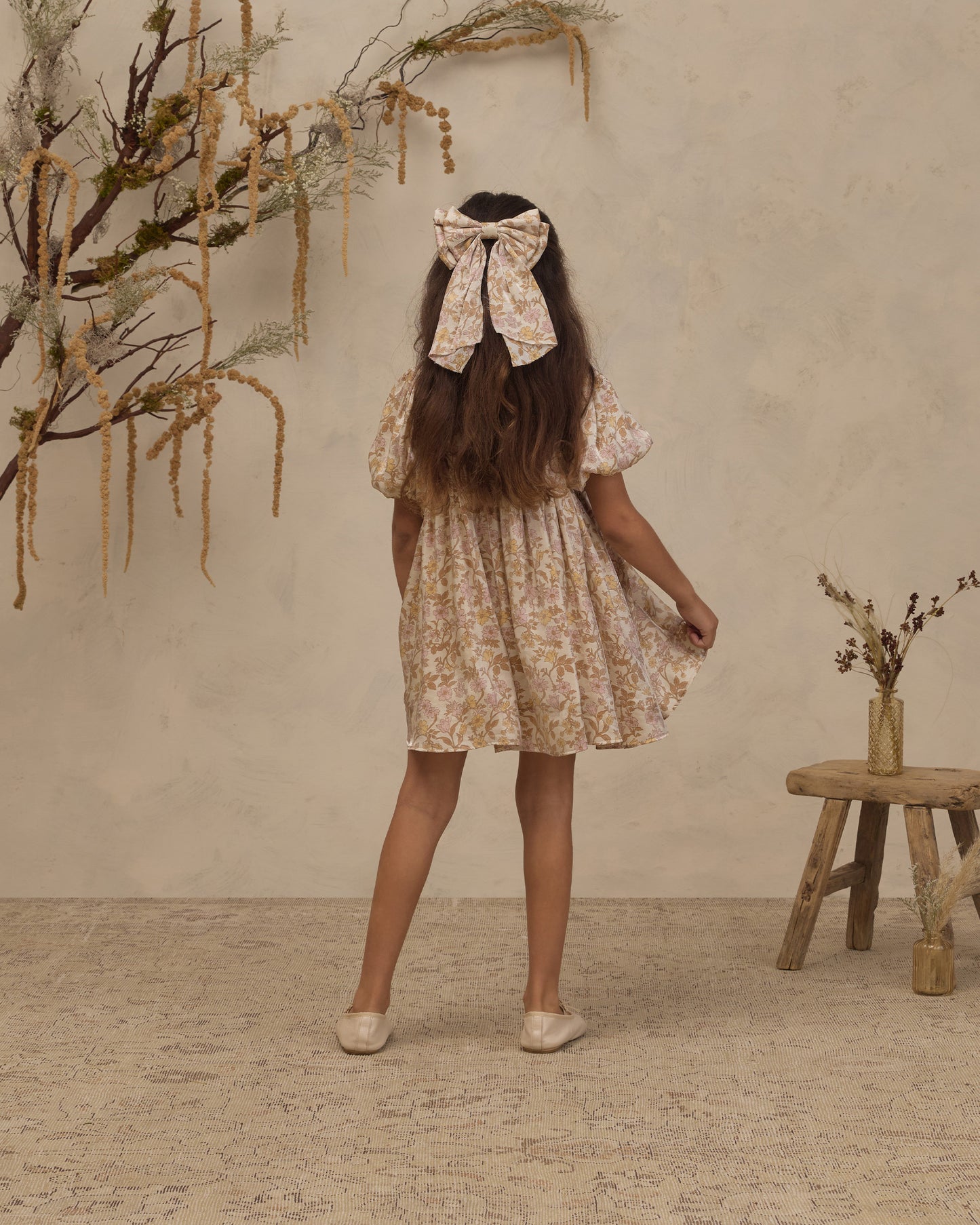 A young girl is shown from behind wearing a short cream-colored floral dress with a matching fabric bow headband. She is standing in a rustic room with a textured beige wall, a small wooden stool, and dried botanical decorations.