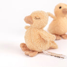 A small plush stuffed duck in a warm beige/tan color, shown from the back with its embroidered closed eyes and a small paper tag attached. Another similar plush duck is softly blurred in the background on a clean white studio surface.