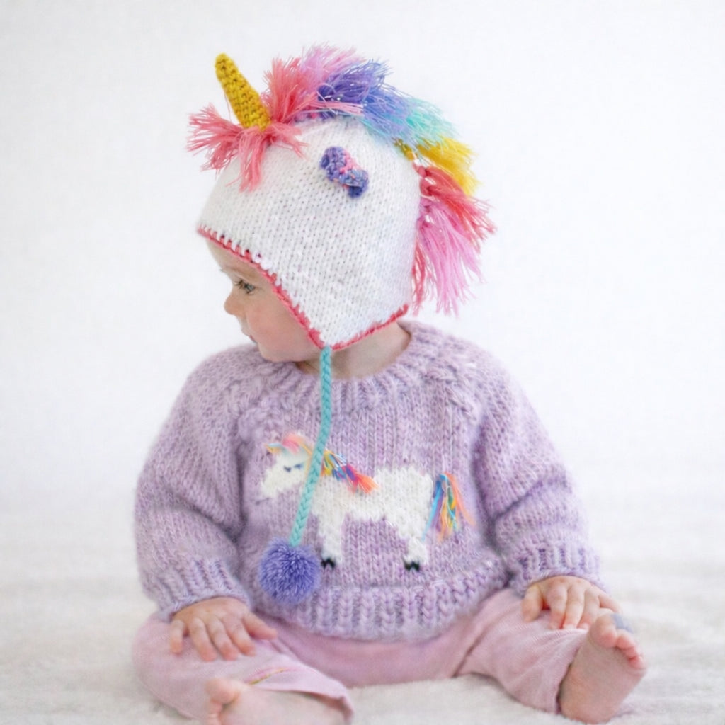 A baby wearing a knitted unicorn-themed hat with a colorful mane and a matching knitted sweater with a unicorn applique, sitting against a plain white background.