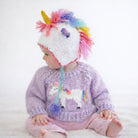 A baby wearing a knitted unicorn-themed hat with a colorful mane and a matching knitted sweater with a unicorn applique, sitting against a plain white background.