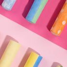 A top-down view of colorful cylindrical items, likely chalk or crayons, arranged on a split pink and light pink background. Visible colors include blue, green, orange with white polka dots, yellow, and light blue.