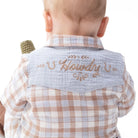 Back view of a baby wearing a light blue muslin neck gaiter with 'Howdy' embroidered in tan script, layered over a beige and white checkered shirt.